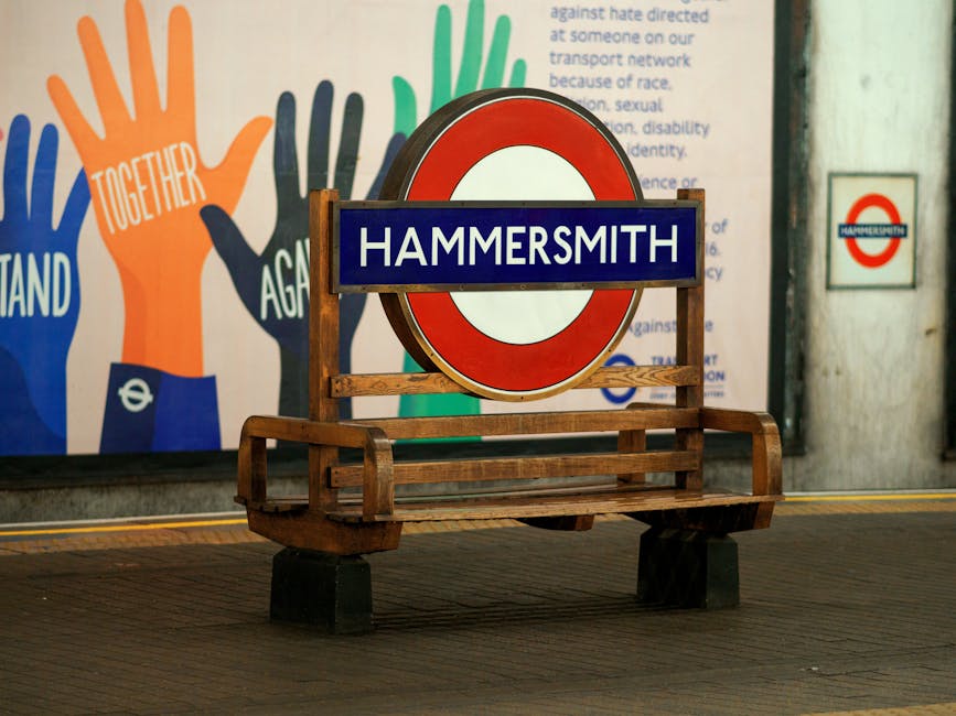 A wooden bench with a slatted seat and backrest is positioned in front of a large, round red and white London Underground 'Hammersmith' station sign mounted on a wooden frame. To the right, there is a small, square sign with the same 'Hammersmith' logo attached to a concrete wall. In the background, a colorful mural featuring multiple stylized handprints in shades of blue, orange, green, and black, with one handprint containing the word 'TOGETHER' in white capital letters, is partially visible. The scene is set on a pavement area with a tactile yellow line running along the edge, suggesting a public transportation environment. The overall setting appears well-lit, with a neutral and professional tone, reflecting an urban station context linked to waste management facilities or public service locations, aligning with the themes of rubbish removal and alternative waste handling associated with independent collection services such as Waste Disposal Hammersmith.