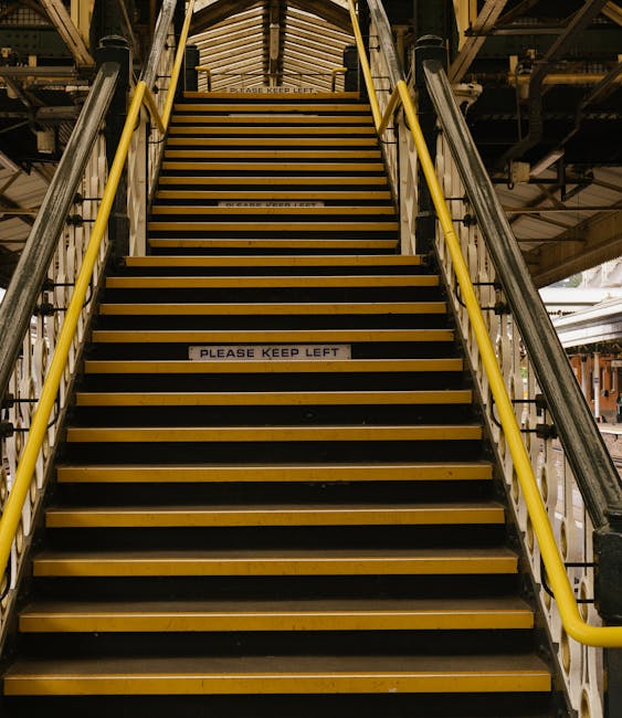 A set of industrial stairs with black and yellow striped treads leading upward within a factory or warehouse environment. The stairs are flanked by yellow handrails on both sides, attached to metallic support structures. Visible parts of the surrounding area include metal beams, supports, and flooring, indicating a utilitarian setting. In the middle of the staircase, a white sign with black text reads 'PLEASE KEEP LEFT,' emphasizing directional guidance for pedestrian flow. The lighting is artificial, casting a neutral tone over the scene, highlighting the industrial materials and textures of the metal stairs and supports. This image suggests an environment where efficient movement and safety protocols are in place, aligning with themes of controlled access and practical waste or materials handling typical in industrial or large-scale waste disposal facilities, such as those operated by Waste Disposal Hammersmith. The straightforward visual presentation underscores the importance of safe accessibility measures in environments where rubbish removal and on-site clearance are conducted, reinforcing the relevance of independent waste collection services within such settings.
