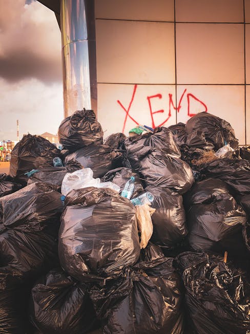 A large pile of black garbage bags filled with waste situated outdoors against a tiled wall with beige and light brown tiles. Several of the bags are tied at the top, some appear slightly torn or sagging. Among the bags, discarded plastic bottles and packaging are visible, scattered around or partially protruding from the bags. In the background, part of a metallic cylindrical structure is seen on the left side, and a cloudy sky with hints of sunlight is visible above, indicating natural daylight. A red spray-painted graffiti on the tiled wall reads 'XEND' in large, slightly uneven letters. The scene exemplifies an excess of waste accumulation, potentially for private disposal or alternative waste collection, relevant in the context of rubbish removal services available from Waste Disposal Hammersmith.
