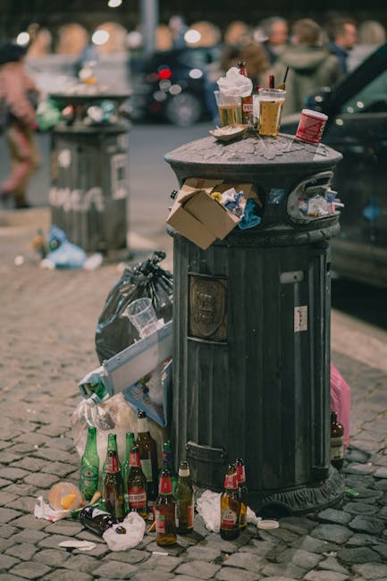 A black public litter bin made of metal and plastic with a domed top, situated on a cobblestone street in an urban environment. The bin is overflowing with various waste items including crumpled paper cups, a cardboard box, and discarded napkins, with additional rubbish such as empty glass bottles, plastic bags, and food wrappers surrounding its base. Several glass bottles with labels are spilled onto the pavement, and crumpled paper and plastic litter are scattered nearby. The background features a blurred crowd of pedestrians, parked cars, and other street furniture, suggesting a busy city scene. The lighting appears natural, indicating daytime, and the overall scene reflects urban waste accumulation that can occur around rubbish collection points when bins are full. The scene represents a typical example of street-level refuse that waste disposal services, such as Waste Disposal Hammersmith, often manage through scheduled or on-demand rubbish removal and waste collection services to prevent overflow and maintain cleanliness.