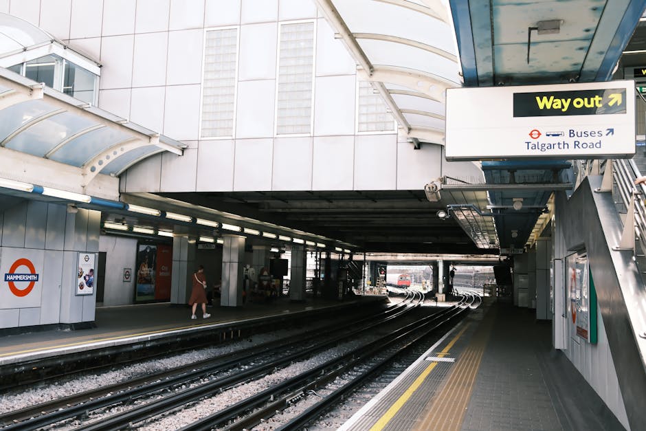 The image depicts an urban train station platform at Hammersmith, with multiple railway tracks running parallel in the foreground. The platform surface is made of dark grey concrete with yellow tactile paving along the edge for safety. To the left, there are a few passengers, including a woman dressed in a red coat walking towards an entrance or exit area, which features white tiled walls adorned with the Hammersmith roundel logo. Above, an arched, semi-transparent roof structure covers the platform, allowing natural light to brighten the area. On the right, a staircase with metallic handrails leads up to a sign indicating the way out, with additional signage directing towards buses and Talgard Road. The environment appears clean and orderly, typical of a busy commuter hub, with the station's design emphasizing durability through metal and glass finishes. Although primarily a passenger transit space, this setting reflects the broader context of urban infrastructure that can contribute to waste management considerations for railway stations and associated services like rubbish removal, managed by providers such as Waste Disposal Hammersmith.