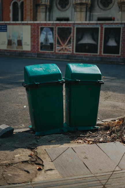 Two green plastic rubbish bins with tightly fitted lids are positioned side by side on a pavement near the edge of a road, with their broad sides facing outward. The bins are made of durable, textured plastic with a matte finish, and each has a horizontal black handle on the front for easy lifting. They are set on a small, flat black base that elevates them slightly above the pavement surface. Surrounding the bins, the sidewalk is partially paved with concrete tiles, some of which have minor cracks and uneven edges, while the adjacent area shows a mixture of dirt and fallen leaves. Behind the bins, a brick-and-mortar building with a decorative architectural style is visible, featuring large windows and a wall painted with partially visible artwork or posters. The road in the background appears clean and is illuminated by natural daylight, suggesting a typical urban environment where private or independent waste disposal might be utilized alongside standard council rubbish collection services. The scene is typical of an area where waste is handled for disposal or recycling, with Waste Disposal Hammersmith potentially offering an alternative waste management option for local residents or businesses.