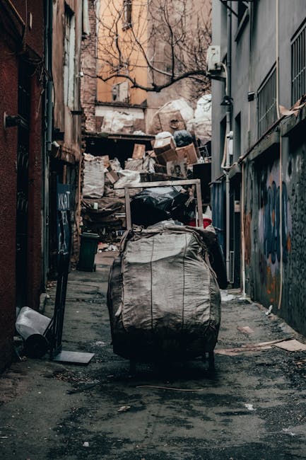 A narrow urban alleyway filled with piles of rubbish and waste materials. In the foreground, a large, grey, industrial-style waste bag is placed on the pavement, with a metal handle and a slightly crinkled surface. Behind it, there are additional scattered waste items, including a black trash bag and various debris, contributing to an untidy scene. Further into the background, an extensive accumulation of cardboard boxes, paper, plastic, and other refuse is stacked haphazardly against a makeshift barrier. The surrounding walls are covered with graffiti and weathered paint, indicating a neglected area, with one side featuring metal fencing and the other showing brick and concrete surfaces. Bare tree branches stretch across the top of the alley, and the lighting appears to be natural, possibly during an overcast day. This scene exemplifies the kind of clutter often managed through private waste collection services, such as those provided by Waste Disposal Hammersmith, for efficient rubbish clearance and site tidy-up, especially in inner-city environments where on-site or independent waste disposal might be necessary.
