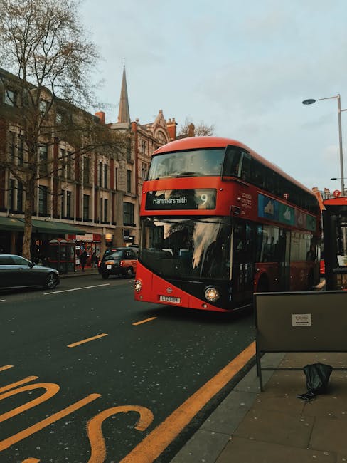 A double-decker bus painted in bright red and black, displaying the destination 'Hammersmith' on the front, is traveling along a city street with multiple lanes. The bus is positioned in the middle of the road, with a slight reflection visible on its curved windshield. The surrounding environment includes a row of tall, multi-storey buildings with a mix of brick and stone facades, some with large windows, lining the street. In the background, a church steeple is visible rising above the rooftops, indicating a historic or ecclesiastical structure nearby. To the left, a few other vehicles such as cars and a van are seen parked or driving along the street. A streetlamp extends from the right side of the image, casting a soft light, and a roadside bench and a black bag are visible on the sidewalk in the foreground. The overall scene is captured during overcast weather, with diffuse natural light illuminating the urban environment, and the image subtly reflects typical city transportation and the infrastructure related to private or alternative waste handling practices, such as rubbish collection, in a London suburb.