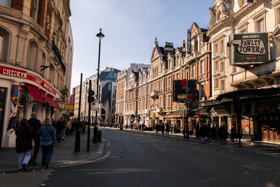 A busy urban street scene showing a mix of historic and modern buildings lining both sides of the road, with pedestrians walking along the wide sidewalk and crossing at a designated crossing point. The buildings feature ornate facades, including decorative window moldings and intricate architectural details, with some displaying signs for theatres and entertainment venues such as 'Fawty Towers' and 'Lyric'. The street is equipped with black street lamps and traffic signals, with a visible bus lane and traffic lights at the corner. The scene is illuminated by natural daylight, casting shadows on the pavement, and the area appears to be a commercial district with shops and theatres. The environment suggests an active city centre setting, typical of a location with historic architecture and vibrant pedestrian activity, relevant to private and independent rubbish removal services operating within urban environments to manage waste from commercial and retail establishments.