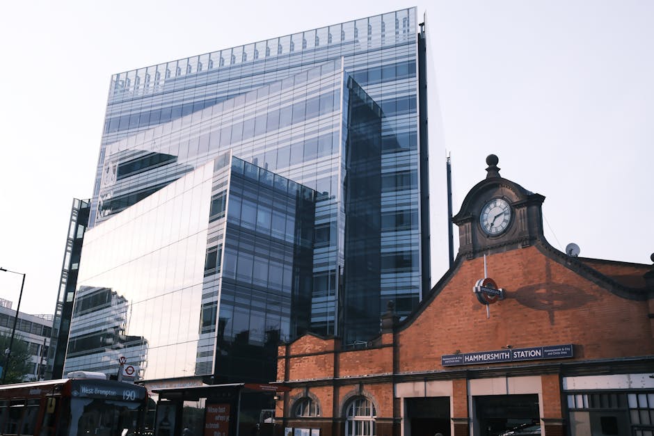 A panoramic view of Hammersmith featuring the modern glass-fronted office building reflecting the sky and surrounding structures, situated behind the historic brick facade of Hammersmith station, which includes a small clock tower and traditional signage. The scene captures the contrast between contemporary architecture and historic station architecture, with the station's brickwork in a warm reddish-brown tone and the clock face prominently displayed. In the foreground, part of a red-brown bus route sign and some street-level advertisements are visible. The environment suggests an urban setting with overcast daylight, and the scene exemplifies an area where independent waste collection or private rubbish removal services could be employed to manage waste in the vicinity of such mixed-use developments, aligning subtly with alternative refuse handling practices typical of urban centres.