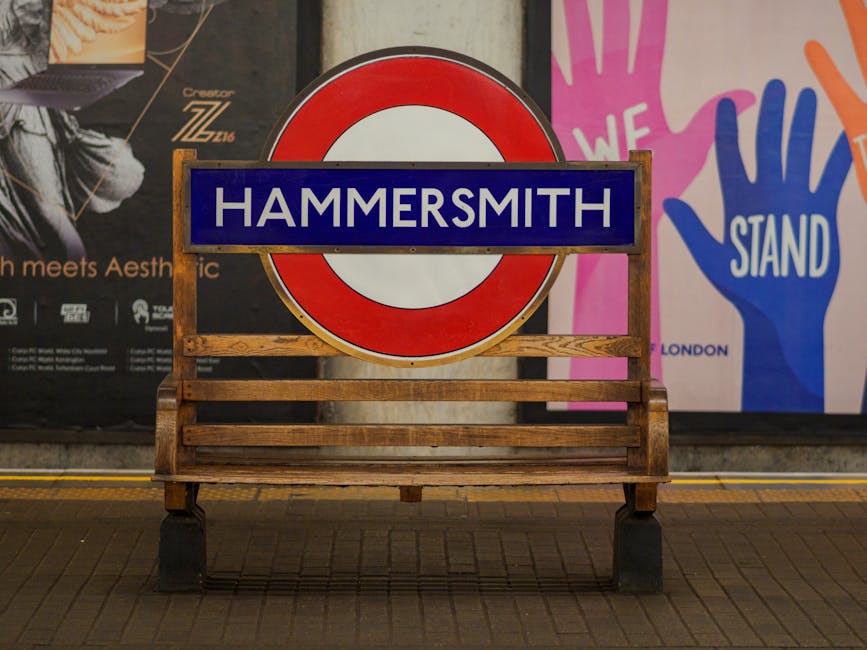 A wooden bench with horizontal slats and a dark metal frame is positioned on a tiled pavement in front of a large red and white circular London Underground sign that prominently displays the word 'Hammersmith' in white letters on a blue background. The background includes a dark wall with partially visible posters and advertising graphics, one of which contains the text 'London' and colorful hand illustrations with the words 'We Stand.' The scene is illuminated by indoor or street lighting, casting slight shadows beneath the bench, and the overall environment suggests a public transportation station or public space area that may be associated with traditional on-site rubbish removal or waste collection practices, with the bench serving as a neutral fixture within the setting. Waste Disposal Hammersmith offers alternative private waste handling services that might be supported by such urban environments.