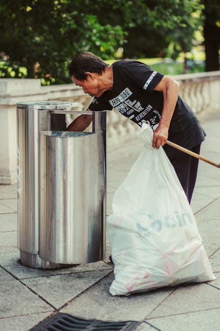 An outdoor scene in an urban setting showing a large pile of mixed household waste and rubbish overflowing from several black, grey, and red wheelie bins on a paved area. The waste includes cardboard boxes, crumpled paper, plastic bags, and discarded packaging. To the left, a grey car is parked behind a metal railing, with its license plate visible. In the background, there is a building with a blue scaffolding structure, windows, and a ground-floor shopfront with yellow and gray signage, indicating a commercial area. Trees are visible along the edge of the scene, and the lighting suggests an overcast day. The image reflects a situation where independent rubbish collection or waste disposal services might be needed, illustrating the importance of efficient rubbish removal in urban environments, and is relevant to services such as those offered by Waste Disposal Hammersmith.
