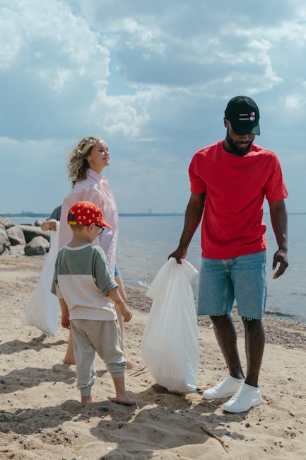A man dressed in a red t-shirt, light denim shorts, white sneakers, and a black cap is standing on a sandy beach, holding a large white plastic bag for rubbish collection. Beside him, a young boy wearing a beige outfit with a red hat decorated with yellow dots is also holding a rubbish bag. In the background, a woman with curly blonde hair, dressed in a light pink or white outfit, stands near the shoreline, facing towards the water and rocks. The scene is set outdoors under a partly cloudy sky, with the sea and a distant horizon visible. The image conveys a scene of outdoor rubbish collection, with the man and children engaging in a beach clean-up activity, matching the context of independent waste disposal and environmental caring. The environment appears natural, with sand, rocks, and calm water, emphasizing the importance of rubbish removal from recreational outdoor spaces as part of waste management activities.